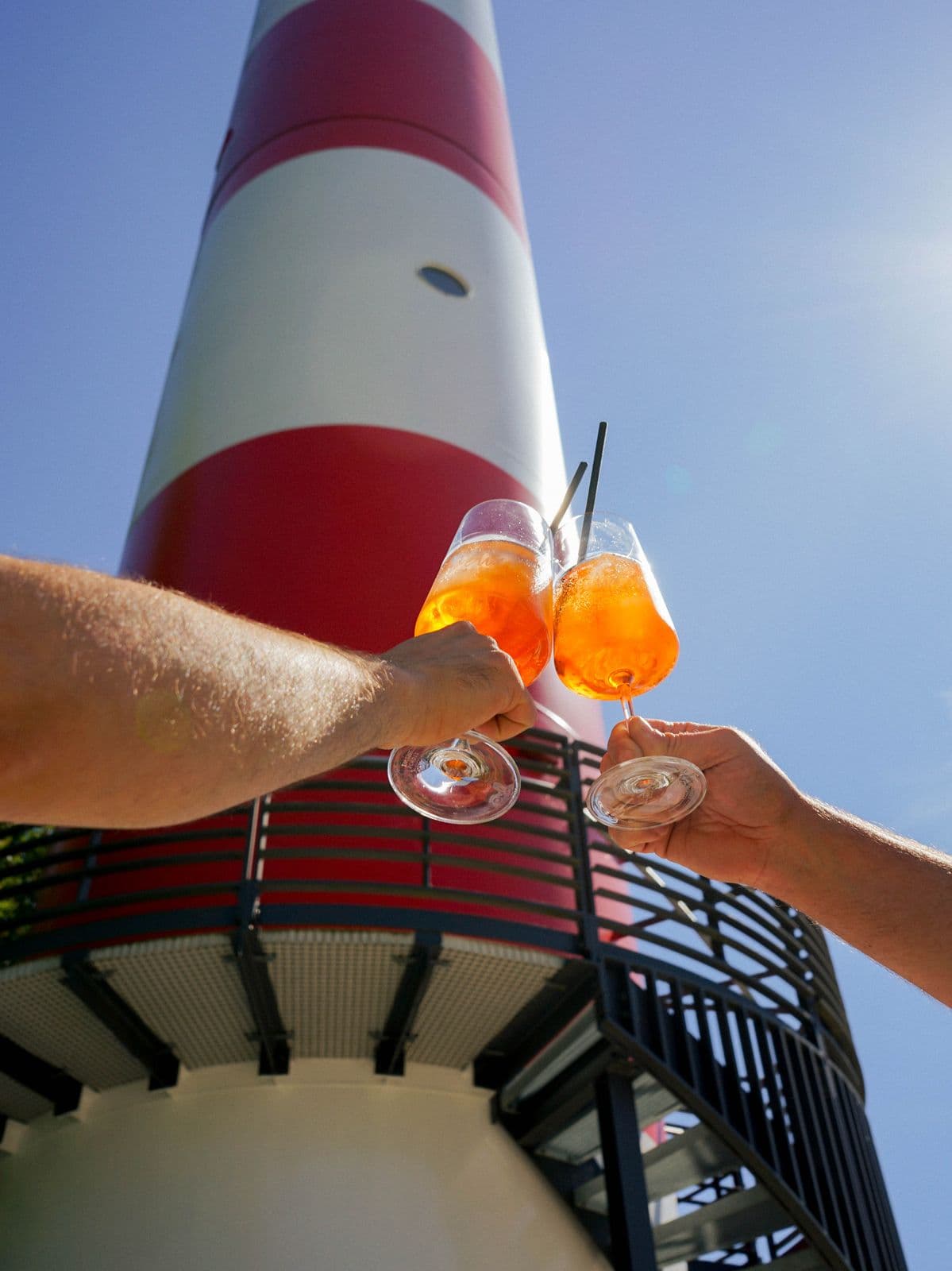 Zwei Gläser Aperol Spritz stoßen vor dem rot-weißen Leuchtturm an